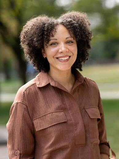 Woman with curly hair smiles outdoors wearing a brown collared shirt.