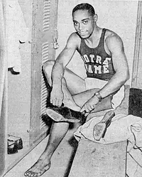A Black basketball player in a Notre Dame uniform puts on his shoes in a locker room.